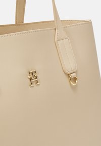 Borsa tote in pelle beige con texture liscia, con emblema dorato e dettagli in metallo. Manici piatti robusti per il trasporto.