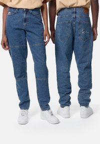 Μπλε τζιν παντελόνι με χαλαρή εφαρμογή, με αντίθετους πάνελ και διπλές ραφές. Λευκά αθλητικά παπούτσια ολοκληρώνουν το σύνολο. Δερμάτινη ετικέτα στη ζώνη.