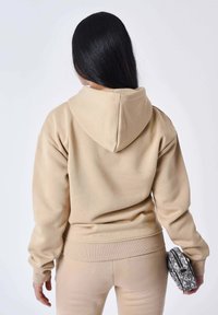 Sudadera con capucha beige hecha de un tejido suave, con puños y dobladillo acanalados. Llevada con pantalones a juego y un bolso gris estampado.