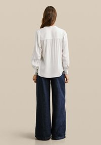 Blusa bianca a maniche lunghe con cucitura arricciata sul retro, abbinata a pantaloni a gamba larga in denim scuro, su uno sfondo neutro.