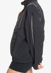 Svart vindjacka med ståkrage, elastisk nederkant och reflekterande sömmar. Tillverkad av lätt och strukturerad tyg.