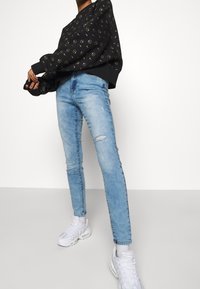 Sweat-shirt noir avec un motif circulaire doré, jean skinny bleu clair avec détails déchirés, et baskets blanches. Look simple et décontracté.