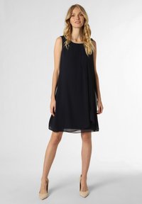 Robe sans manches de couleur bleu marine foncé avec un design fluide, des détails de drapé subtils et un tissu léger. Associée à des escarpins beiges.