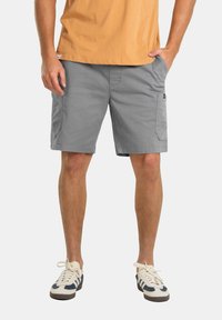 Pantalones cortos de carga grises con bolsillos, material de algodón, corte recto, combinados con zapatillas crema y marino, llevados con una camiseta naranja.