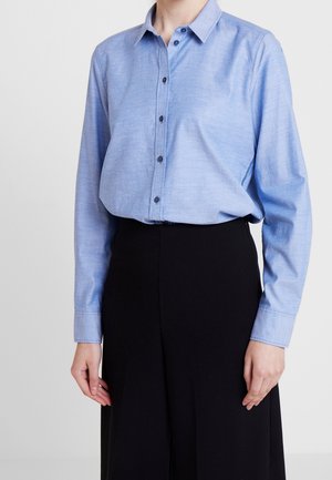 Person iført en lyseblå skjorte med knapper og lange ærmer, stoppet ned i sorte bukser med høj talje, stående mod en ensfarvet lys baggrund.