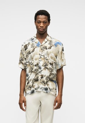 Mężczyzna ubrany w koszulę na krótki rękaw z abstrakcyjnymi wzorami w kolorach czarnym, beżowym i niebieskim, w połączeniu z jasnobeżowymi spodniami.