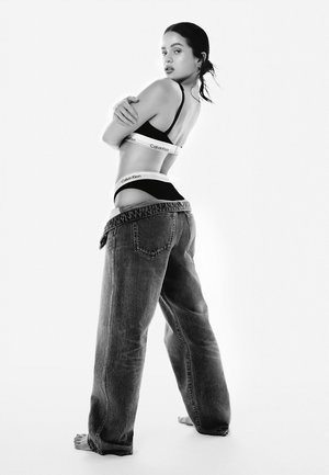Un reggiseno e una fascia Calvin Klein neri visibili con jeans in denim blu a vestibilità morbida, leggermente s saggiati, che rivelano un design liscio e minimalista.