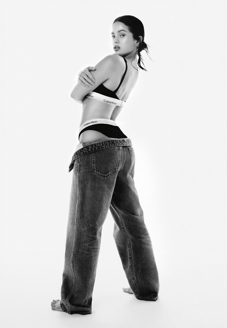 Un soutien-gorge noir Calvin Klein et une ceinture visibles, associés à un jean en denim bleu ample, légèrement sagging, mettant en avant un design lisse et minimaliste.