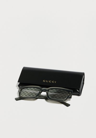 Ορθογώνια γυαλιά ηλίου Gucci με γκρι φακούς με μοτίβο που ακουμπούν μπροστά σε μαύρη θήκη Gucci με χρυσά γράμματα σε λευκό φόντο.