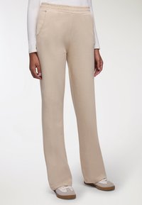 Pantaloni della tuta beige con una vestibilità ampia, vita elasticizzata e tasche. Il tessuto appare morbido e ha una texture liscia; indossati con sneakers bianche casual.