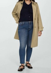 Trench-coat beige porté sur une chemise à boutons bleu marine, associé à un jean skinny bleu et des chaussures plates noires. Le manteau est doté d'une taille cintrée avec ceinture et de boutons décoratifs.