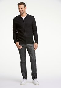 Musta vetoketjullinen neule, jossa on ribsineuleiset hihat ja helma, yhdistetty tummanharmaisiin farkkuihin ja valkoisiin tennareihin, seisoo vaalean valkoista taustaa vasten.