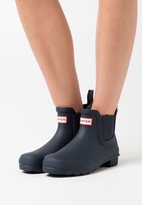 Bottes en caoutchouc navy à la cheville avec des semelles texturées et des panneaux latéraux élastiques. Présente une étiquette de marque rouge et blanche à l'avant.