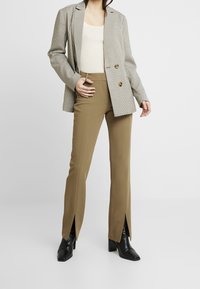 Blazer à carreaux avec boutons dorés, associé à un pantalon vert olive avec des fentes sur les côtés. Des bottines noires à la cheville complètent la tenue.