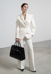 Blazer blanc sur mesure avec un pantalon assorti, doté d'une ceinture. Sac à main noir texturé avec deux poignées. Le modèle se tient devant un fond clair.