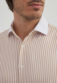 Chemise rayée avec des rayures verticales blanches et beiges, un col blanc, et deux boutons au niveau du cou. Le tissu semble léger et texturé.