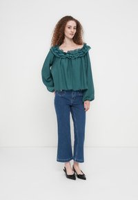 Τόπ με ώμους έξω σε πετρόλ χρώμα, με βολάν στον λαιμό, μακριά μανίκια και φαρδιά εφαρμογή, συνδυασμένο με cropped μπλε τζιν και μαύρα παπούτσια.