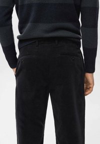 Pantaloni di velluto a coste neri con due tasche posteriori, chiusura a bottone e un design a gamba dritta. Il tessuto ha una superficie morbida e testurizzata.