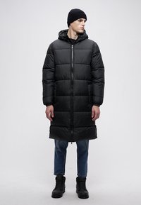 Zwarte gewatteerde lange jas met ritssluiting, afneembare capuchon en zijzakken, gecombineerd met blauwe spijkerbroek en zwarte veterlaarzen.