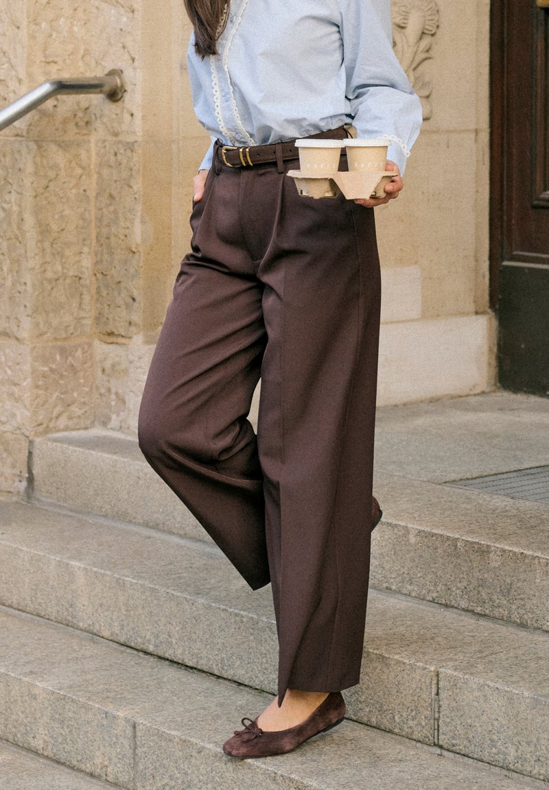 Personne debout sur des marches en pierre tenant un porte-quatre-tasses de café, portant un pantalon marron, une chemise bleue et des ballerines marron foncé.