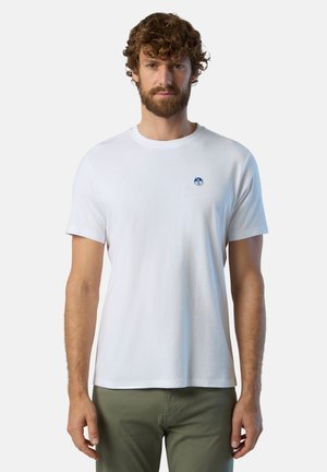 T-shirt bianco in cotone con scollo rotondo, maniche corte e un piccolo logo ricamato blu sul petto, abbinato a pantaloni verde oliva.