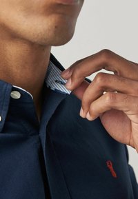 Chemise boutonnée bleu marine avec un col rayé contrastant; présente un logo de homard rouge brodé sur la poitrine et est fabriquée en coton.