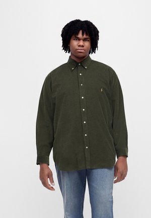 Jeune homme aux cheveux courts et bouclés portant une chemise verte foncée à manches longues boutonnée et un jean bleu, debout devant un fond blanc uni.