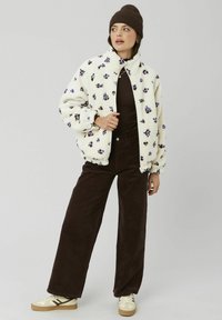 Veste en fausse fourrure crème avec broderies florales violettes ; fermeture éclair, col montant et coupe décontractée. Associée à un pantalon en velours côtelé marron et des baskets beiges.