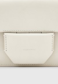 Beige lædertaske med en geometrisk klaplukning og diskret syninger. Har et præget "ALLSAINTS"-logo på forsiden. Glat tekstur.