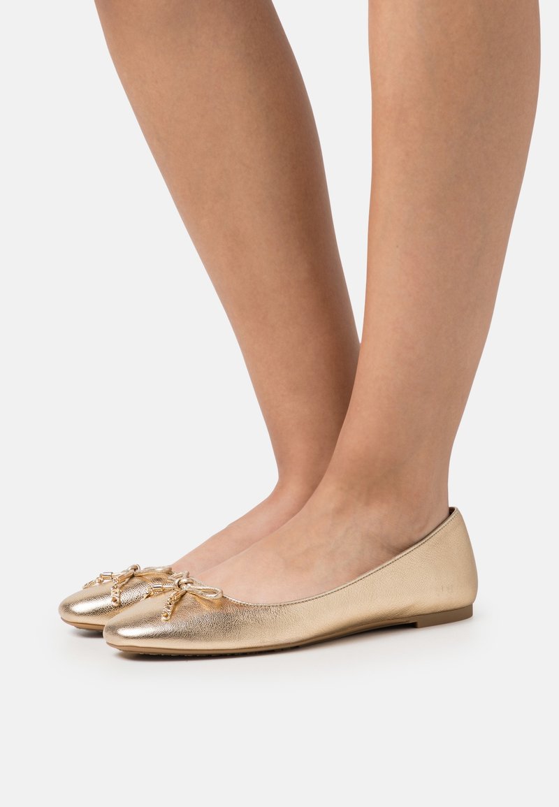 Jambes portant des ballerines dorées métalliques avec un petit détail chaîne et nœud sur le dessus, sur un fond clair uni.