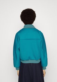 Tjurkos texturerad jacka med en strukturerad krage, elastiska ärmslut och midja, samt en horisontell sömdetalj över ryggen.