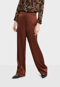 Pantalons larges en satin marron avec une texture lisse, dotés d'une taille haute et d'un léger brillant, associés à des chaussures noires à bout pointu.