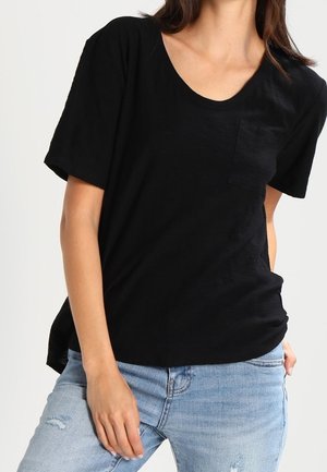 Kobieta w czarnym, krótkim rękawem z kieszeniami t-shircie i jasnoniebieskich podartych dżinsach, pozująca z jedną ręką blisko uda.