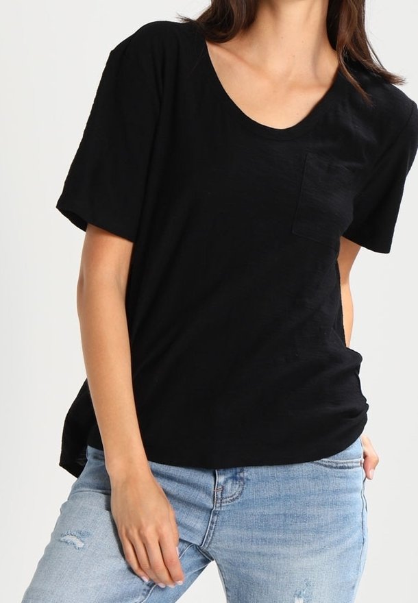 Femme portant un t-shirt noir à manches courtes avec poche et un jean bleu clair déchiré, posant avec une main près de sa cuisse.