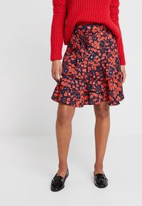 Hame floral dalam warna navy dengan bunga merah dan oranye, tepi berbulu, dan ikatan samping. Dipadukan dengan sweater rajut merah dan sepatu loafer hitam.