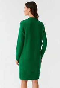 Robe tricotée verte avec un ourlet et des poignets côtelés. Présente des accents de boutons à l'épaule. Texture douce, design mi-long.