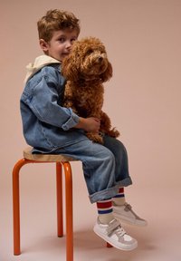 Un enfant en veste et pantalon en denim est assis sur un tabouret orange, tenant un chien brun aux cheveux bouclés. Il porte des chaussures blanches avec des accents gris et des chaussettes rayées.