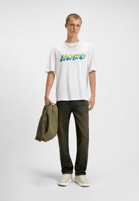 Camiseta blanca con un gráfico multicolor de "HUGO", combinada con jeans oscuros y zapatillas beige. Sostiene una sudadera verde con la mano izquierda.