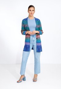 Colorful, patterned cardigan with long sleeves and open front. Worn over a ribbed light blue top, paired with high-waisted light wash jeans and blue heels.