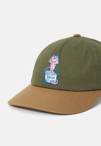 Olivengrøn kasket med en tan skygge, der har en broderet blå og pink tegneseriefigur på forsiden med teksten "HUF" nedenunder.