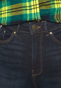 Jeans de mezclilla de un azul oscuro con un ajuste ceñido, con costuras de cobre y un botón de latón con la inscripción "ONLY". Combinado con una camisa de franela a cuadros en verde y amarillo.