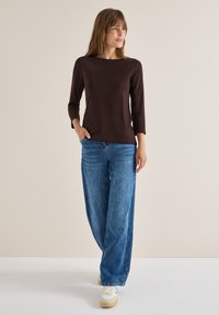 Braunes Langarmoberteil kombiniert mit weit geschnittenen blauen Jeans. Model trägt Sneaker und steht vor einem neutralen Hintergrund.