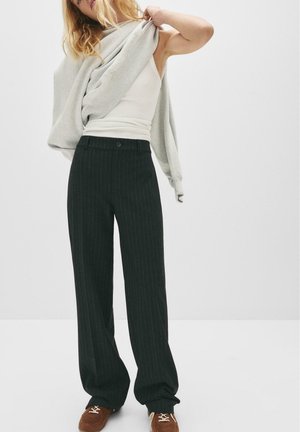 Szary sweter zarzucony na ramiona, biały top na ramiączkach pod spodem i czarne szerokie spodnie w paski. Brązowe buty dopełniają ten zestaw.