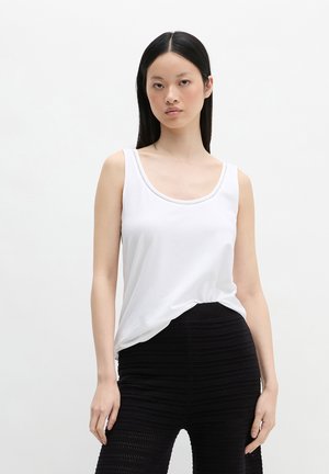 Vrouw met stijl zwart haar die een witte mouwloze top en zwarte getextureerde hoge taille broek draagt, staand tegen een witte achtergrond.
