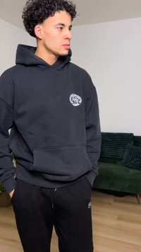 Black hoodie with a front pocket and circular logo on the chest, paired with black sweatpants. Material appears soft and casual.