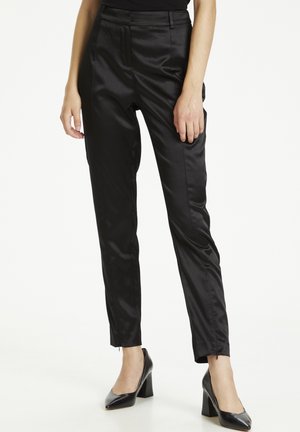 Woman wearing fitted black satin trousers with ankle zippers and black high-heeled pointed-toe shoes, standing against a white background.