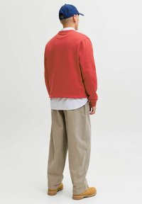 Czerwona bluza z długim rękawem, nałożona na biały pasek koszuli, sparowana z luźnymi beżowymi spodniami i brązowymi butami, widok z tyłu.