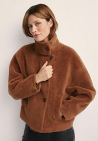 Brązowa polarowa kurtka z wysokim kołnierzem, z zapięciem na guziki i bocznymi kieszeniami; miękka faktura, luźny fason i casualowy design.