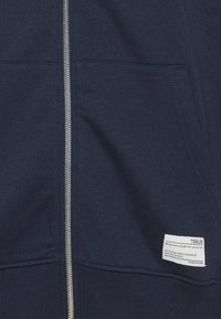 Navy plava zip-up dukserica s prednjim džepom, srebrnim zatvaračem i bijelom pravokutnom oznakom od tkanine ušivenom na donjem desnom dijelu džepa.