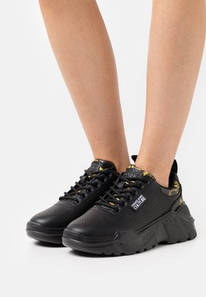 Baskets noires avec un design perforé, semelle épaisse et accents floraux jaunes. Dotées de lacets, d'un col rembourré et d'un logo sur la languette.